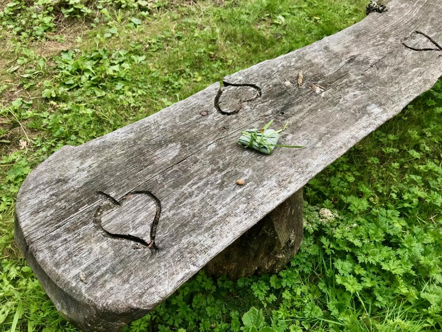 A rustic bench made from a plank with hearts carved into it and a tiny object wrapped in a leaf on top